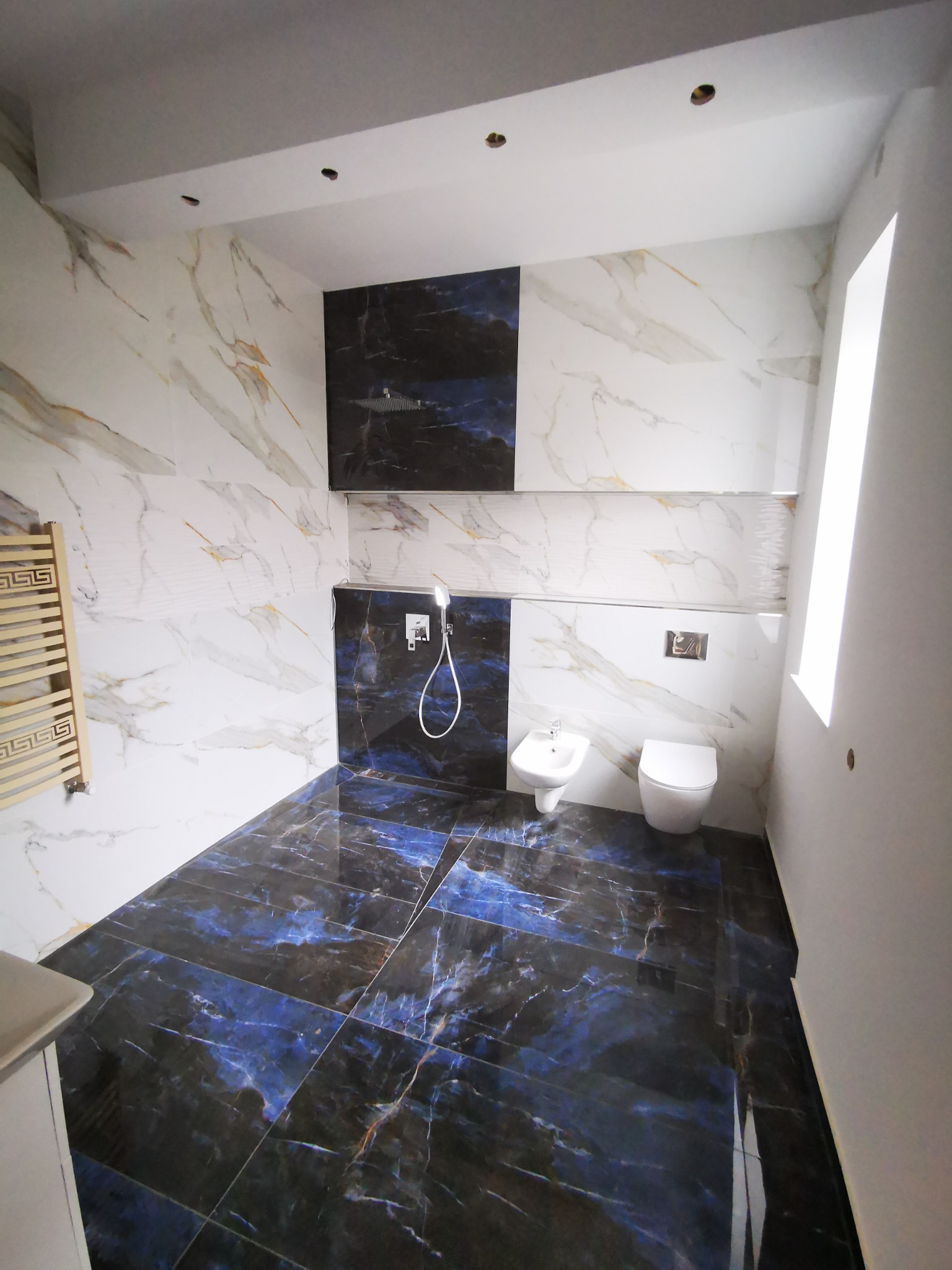 Wykończona łazienka z płytkami imitującymi marmur w odcieniach bieli i granatu, widoczna toaleta, bidet, grzejnik drabinkowy z greckim wzorem, armatura prysznicowa i okno.