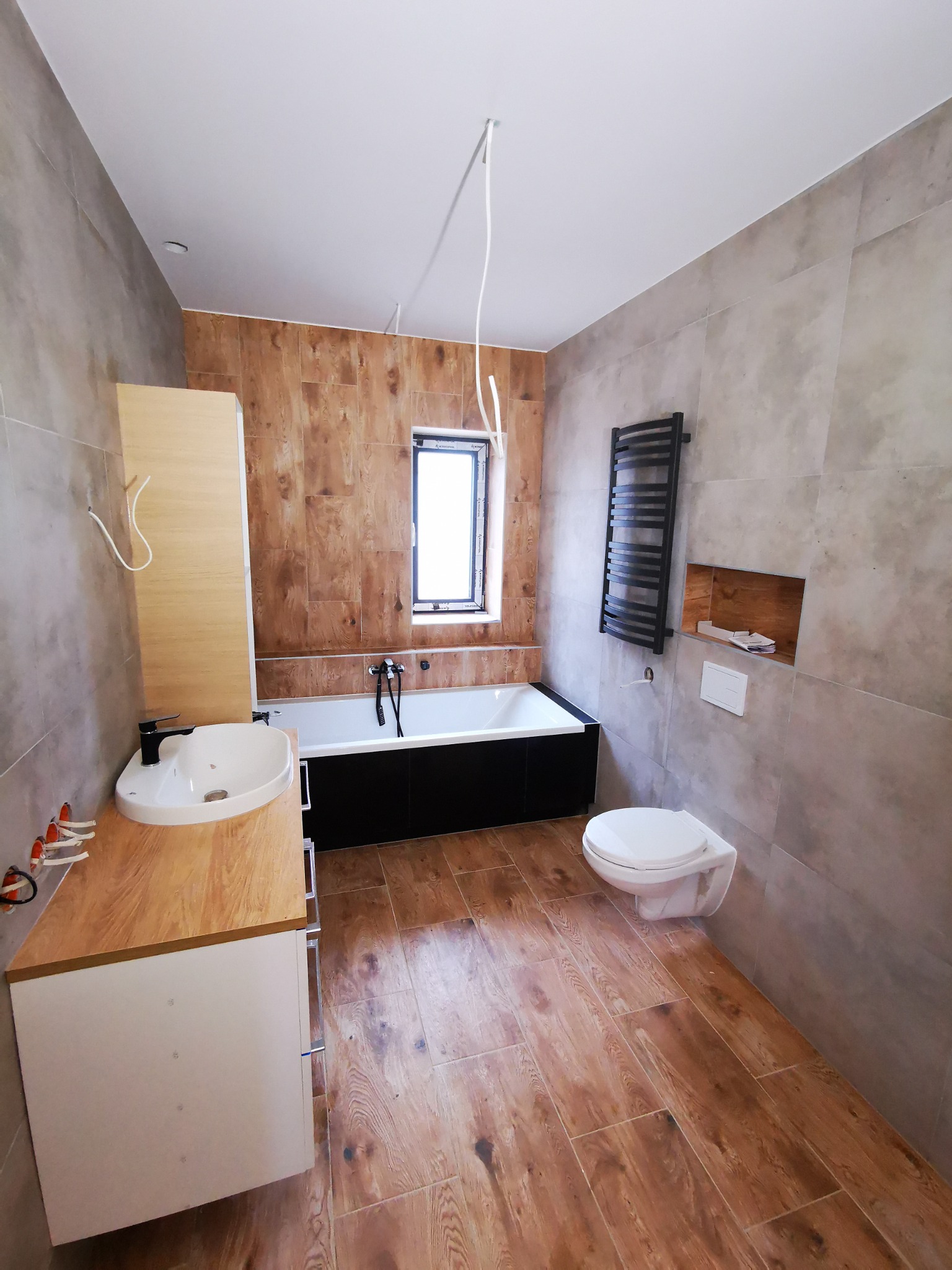 Nowoczesna łazienka z drewnopodobnymi płytkami na podłodze i ścianie, czarna wanna obudowana, umywalka nablatowa na drewnianym blacie, grzejnik drabinkowy i wisząca toaleta. Widoczne wystające...