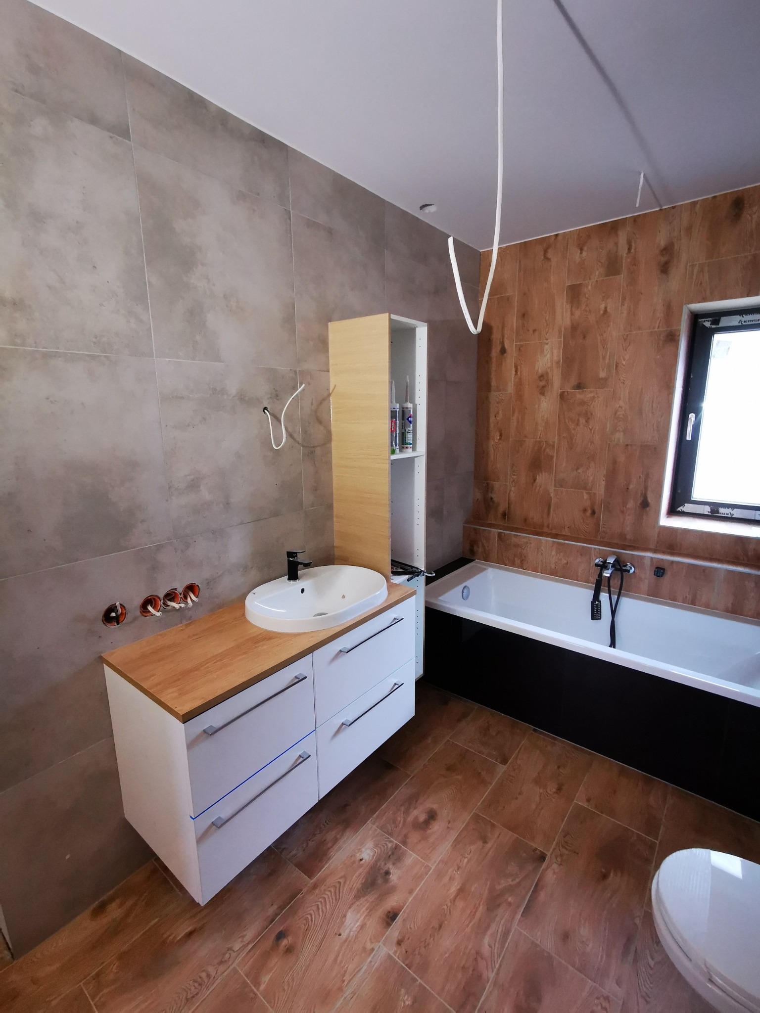 Nowoczesna łazienka w trakcie wykańczania: brązowe płytki imitujące drewno na podłodze i ścianie przy wannie, szare płytki na ścianie z umywalką nablatową na białej szafce z drewnianym blatem...