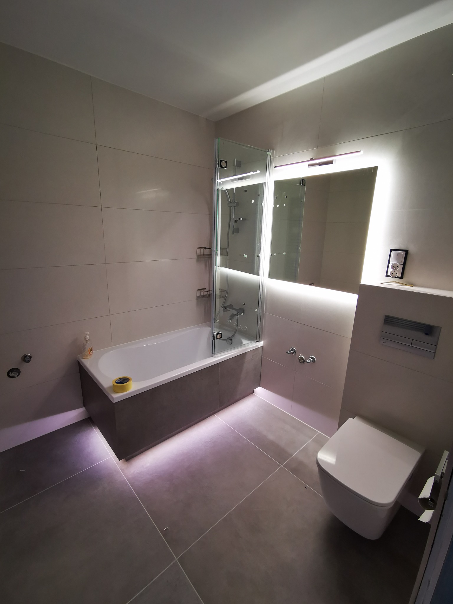 Nowoczesna łazienka z podświetlanym lustrem, kabiną prysznicową nad wanną, wiszącą toaletą i szarą glazurą.