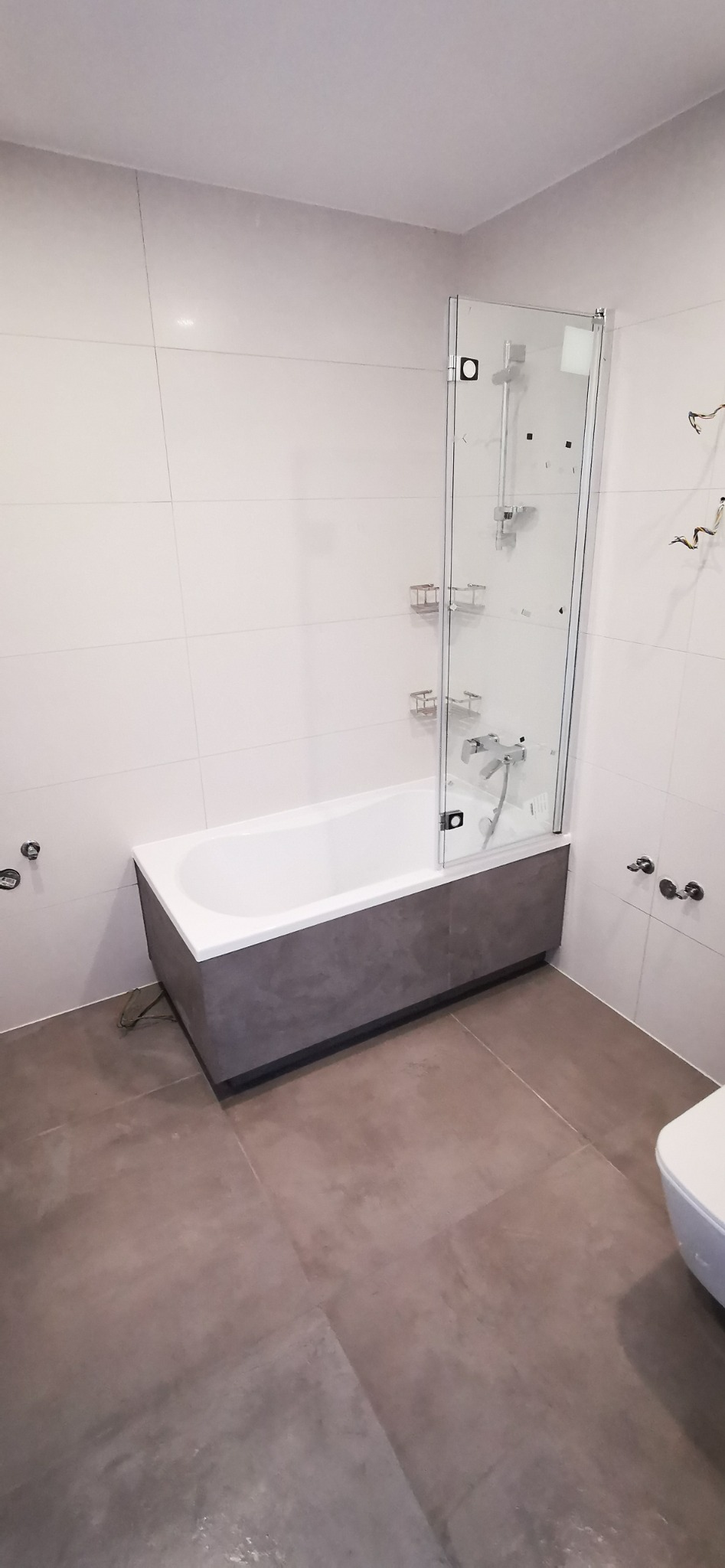 Nowoczesna łazienka z białą wanną obudowaną szarą mozaiką, przeszklonym parawanem nawannowym z półkami, szarą podłogą i białymi płytkami ściennymi, widoczne wystające przewody elektryczne.