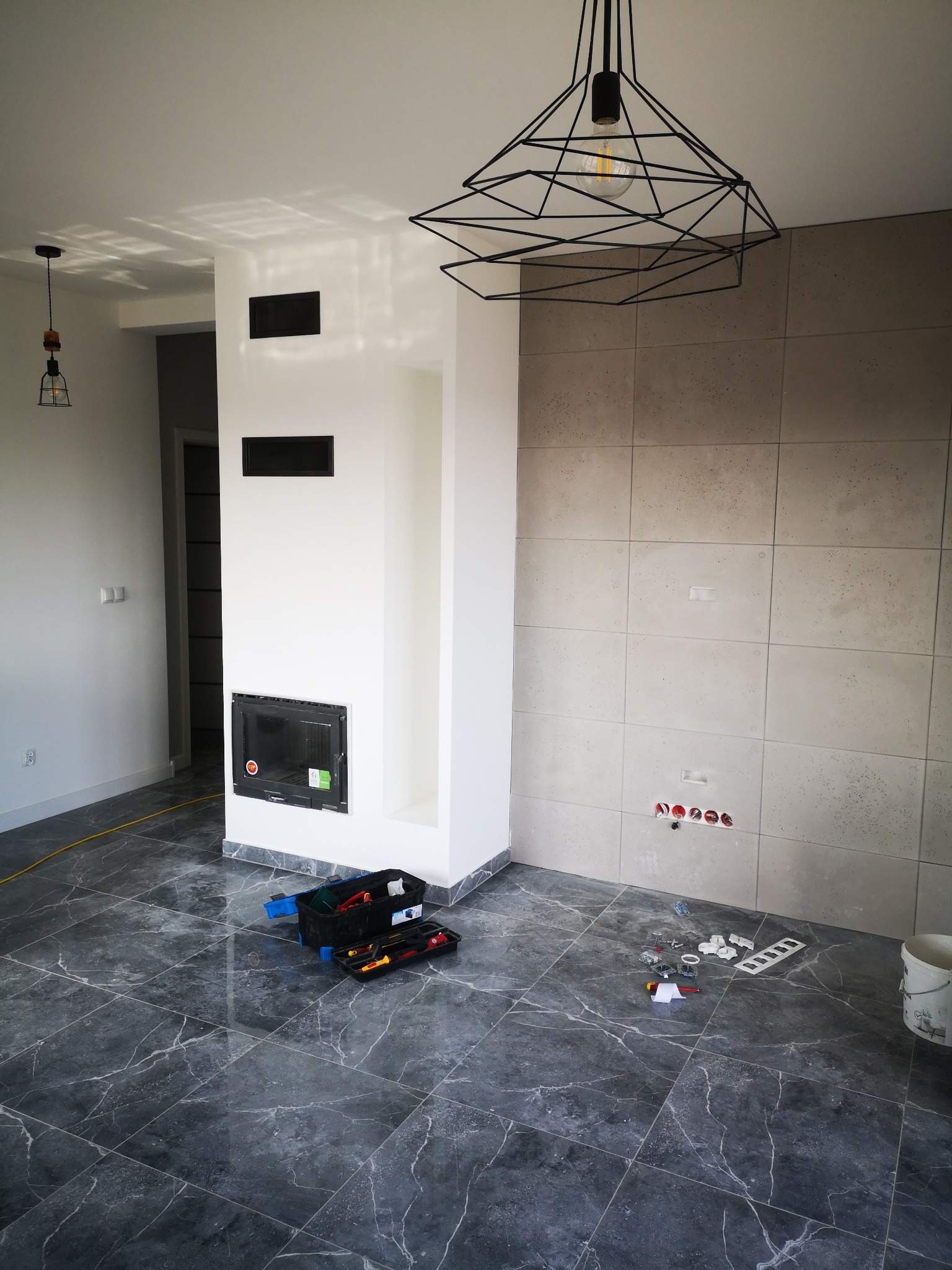 Wnętrze w trakcie wykańczania: szara marmurowa podłoga, ściana wyłożona płytkami imitującymi beton, zabudowany kominek z czarnymi wlotami powietrza, widoczne gniazdka elektryczne, skrzynka...