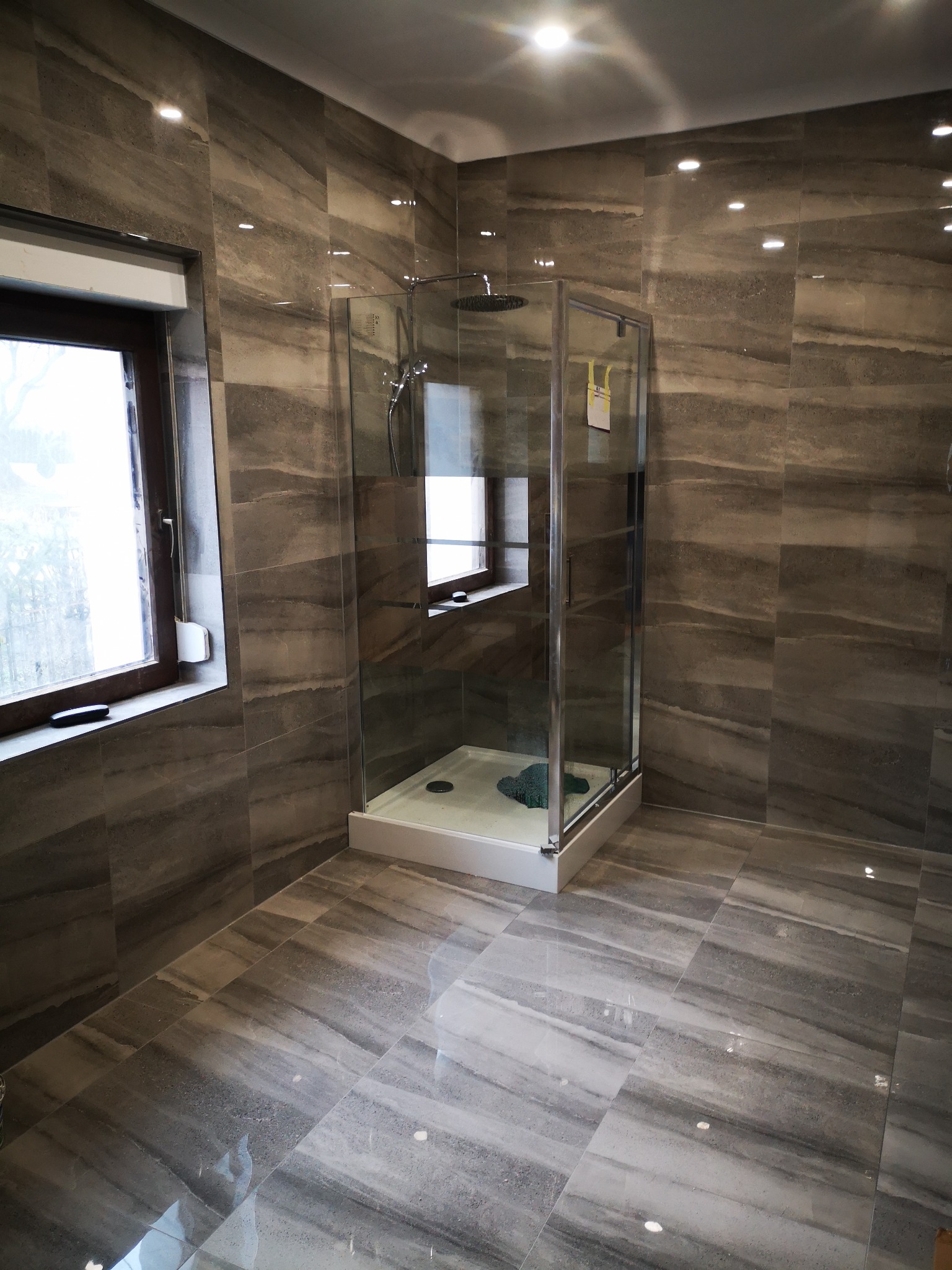 Nowoczesna łazienka z kabiną prysznicową typu walk-in, wykończona płytkami imitującymi marmur w odcieniach szarości, z oknem w ciemnej ramie i punktowym oświetleniem sufitowym.