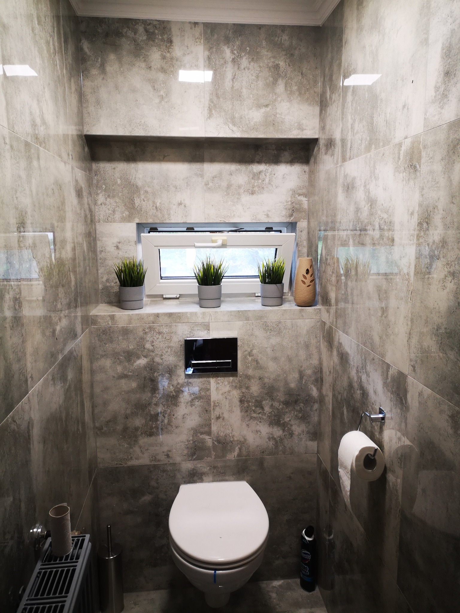 Mała łazienka wykończona szarymi płytkami imitującymi kamień, z podwieszaną toaletą, wąskim oknem z trzema doniczkami z zielonymi roślinami i odświeżaczem powietrza na półce, oraz grzejnikiem...