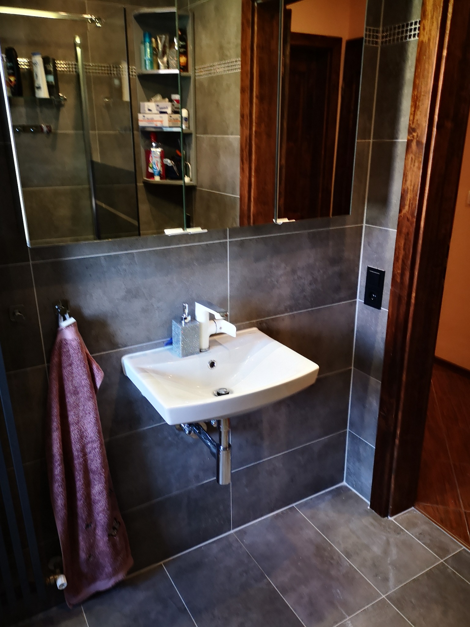 Umywalka nablatowa z baterią i dozownikiem mydła, szara płytka na ścianach, ręcznik na grzejniku drabinkowym w wyremontowanej łazience.