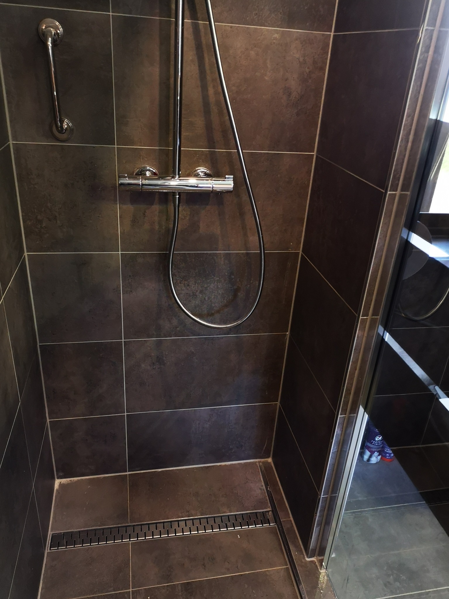 Nowoczesna kabina prysznicowa z ciemnymi, matowymi płytkami, chromowaną armaturą i odpływem liniowym w podłodze.