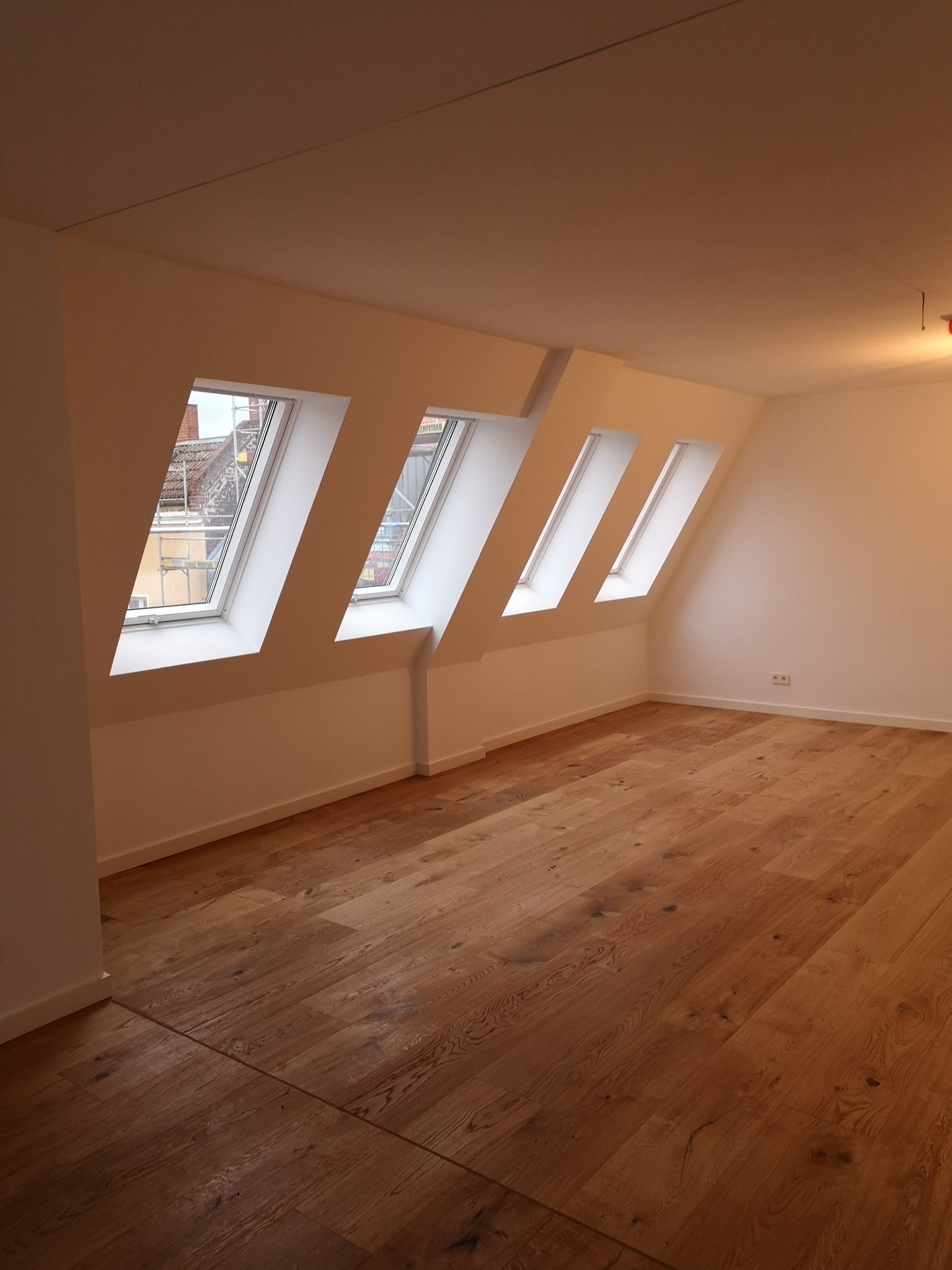 Poddasze z jasnymi ścianami, drewnianą podłogą i czterema oknami dachowymi w ukośnej ścianie, widok na rusztowania za oknem.