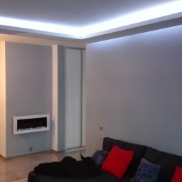 Baufirma Orczykowski - Szare wnętrze pokoju z podświetlanym sufitem napinanym LED, białą ścianą z cegły i nowoczesnym kominkiem wnękowym.