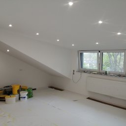 Baufirma Orczykowski - Poddasze po malowaniu na biało, z widocznymi punktowymi lampami w suficie, zabezpieczoną podłogą folią malarską, wiadrami z farbą i narzędziami malarskimi przy ścianie szczytowej oraz zakrytym...