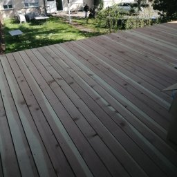 Baufirma Orczykowski - Nowo ułożona drewniana podłoga tarasu z widocznymi śladami po obróbce i impregnacji, na tle budynku mieszkalnego z zielonym trawnikiem i drzewami.