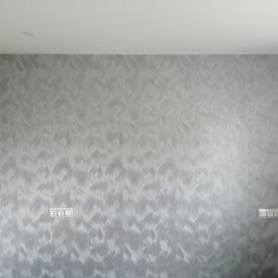 Baufirma Orczykowski - Ściana pomalowana dekoracyjną farbą w odcieniach szarości z efektem falowania, widoczne dwa gniazdka elektryczne i fragment białego sufitu z wbudowanym oświetleniem.