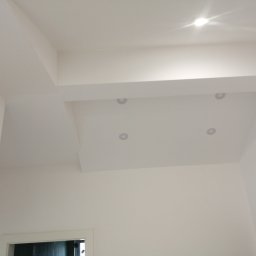 Baufirma Orczykowski - Nowoczesny, biały sufit podwieszany z wbudowanymi punktowymi źródłami światła, widoczne fragmenty ścian i ościeżnica drzwi.