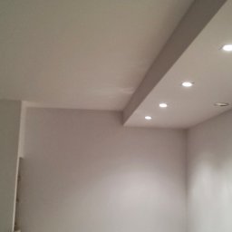 Baufirma Orczykowski - Biały sufit z zamontowanymi punktowymi lampami LED w rzędzie, widoczny fragment ściany z okrągłym głośnikiem.