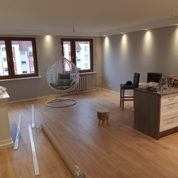Baufirma Orczykowski - Jasne wnętrze mieszkania w trakcie remontu z drewnianą podłogą, widoczne profile i zwinięty materiał, prawdopodobnie do montażu sufitu napinanego, oraz wiszące przewody elektryczne.