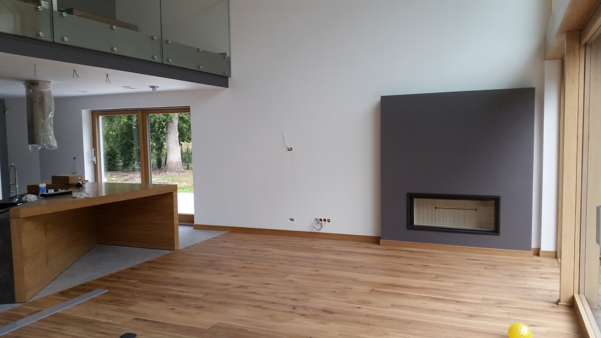 Jasny parkiet ułożony w przestronnym salonie z szarą ścianą kominkową i widokiem na ogród przez duże okno; fragment wyspy kuchennej z blatem z jasnego drewna.