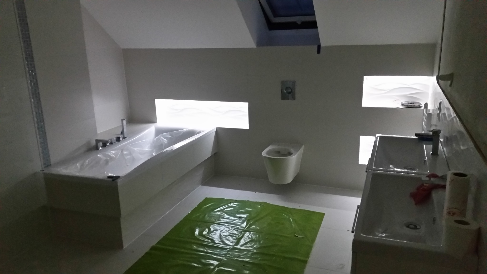 Nowoczesna łazienka w trakcie remontu: wanna zabezpieczona folią, podłoga przykryta zieloną plandeką, umywalki z bateriami, toaleta wisząca, dekoracyjne wnęki z oświetleniem.