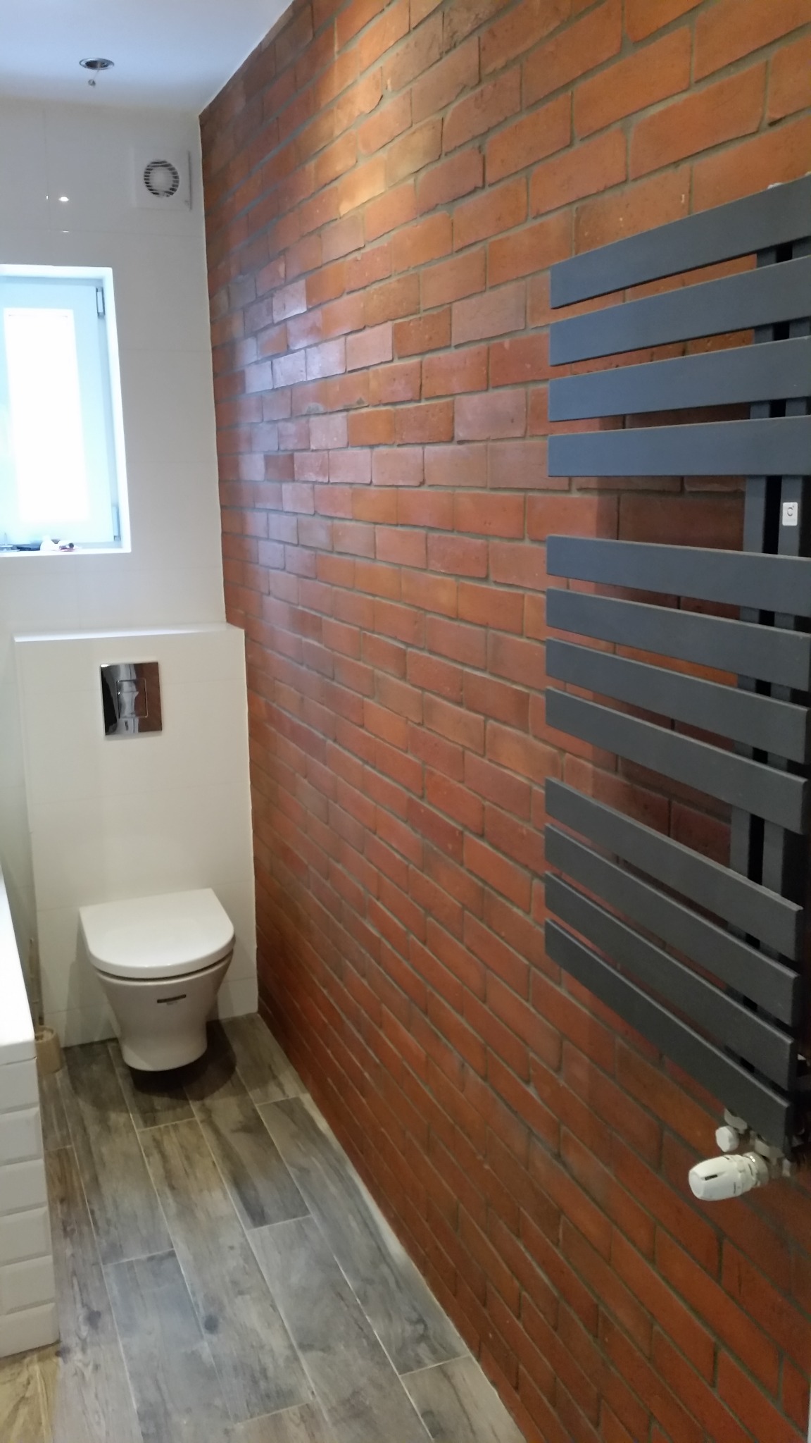Wykończona łazienka z czerwoną ścianą z cegły, szarym grzejnikiem drabinkowym i toaletą podwieszaną na białej ściance, podłoga wyłożona panelami imitującymi drewno.