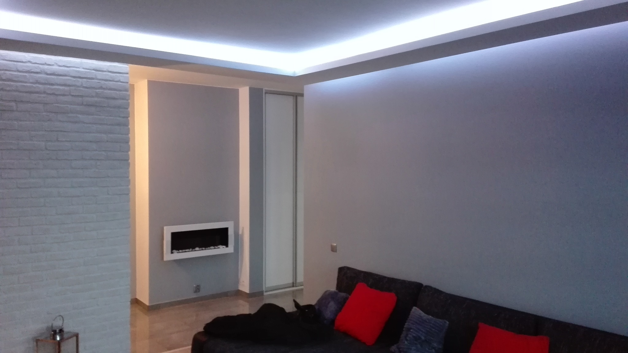 Szare wnętrze pokoju z podświetlanym sufitem napinanym LED, białą ścianą z cegły i nowoczesnym kominkiem wnękowym.