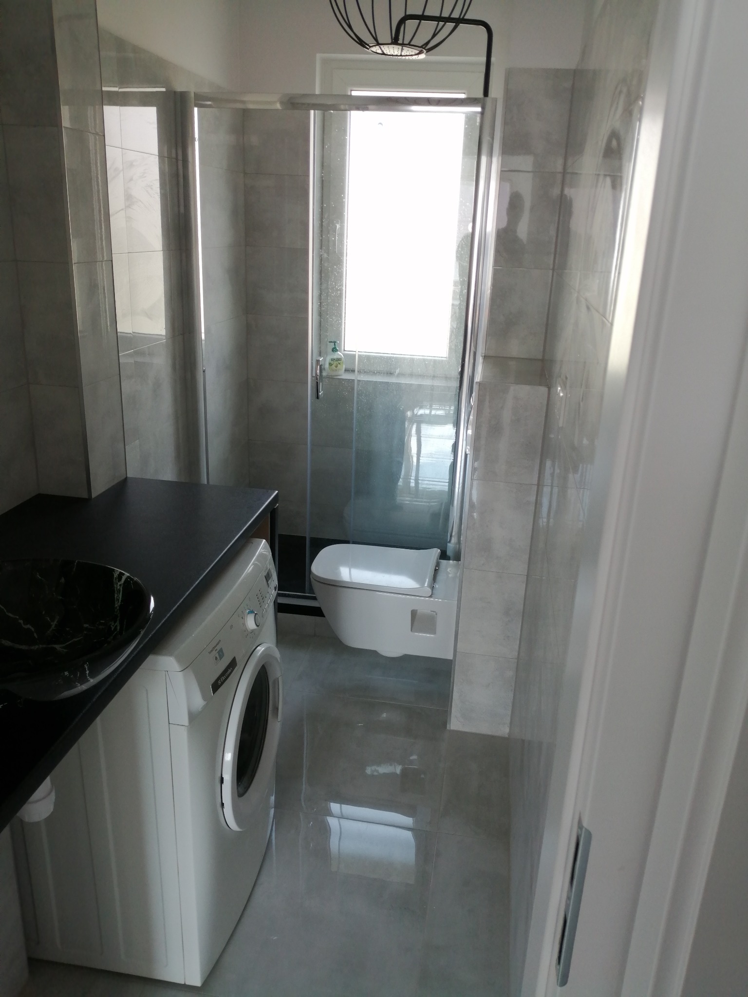 Nowoczesna łazienka z szarymi płytkami, kabiną prysznicową ze szklanymi drzwiami, umywalką w czarnym marmurze i pralką Electrolux pod blatem.