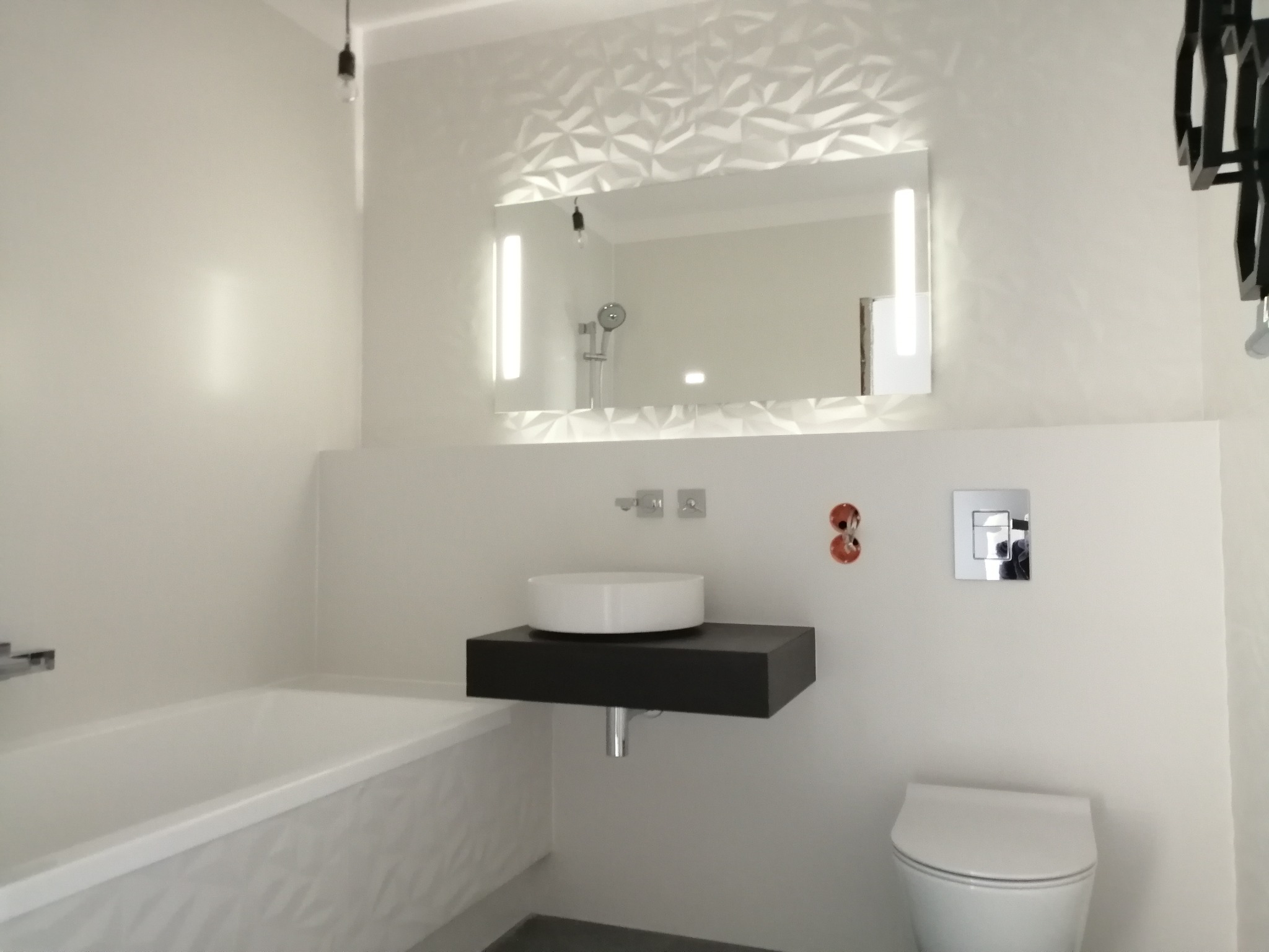 Nowoczesna łazienka w jasnych barwach z geometrycznym wzorem na ścianie, podświetlanym lustrem, okrągłą umywalką na czarnym blacie i wiszącą toaletą.