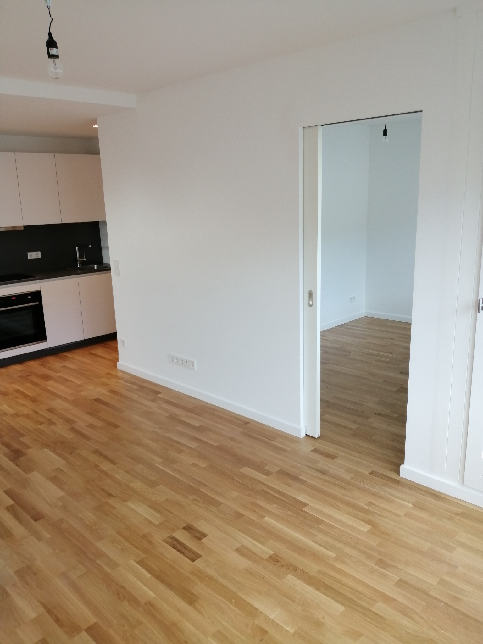 Jasny parkiet ułożony w nowym mieszkaniu z widoczną kuchnią z białymi szafkami i czarnym panelem nad blatem oraz otwartym przejściem do kolejnego pomieszczenia.