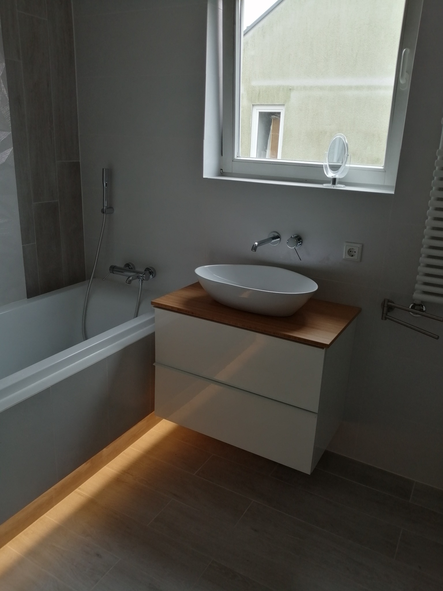 Nowoczesna łazienka z wiszącą szafką pod umywalkę z blatem z jasnego drewna, owalna umywalka nablatowa, szara glazura w strefie wanny, okno z widokiem na budynek i małe lusterko na parapecie.