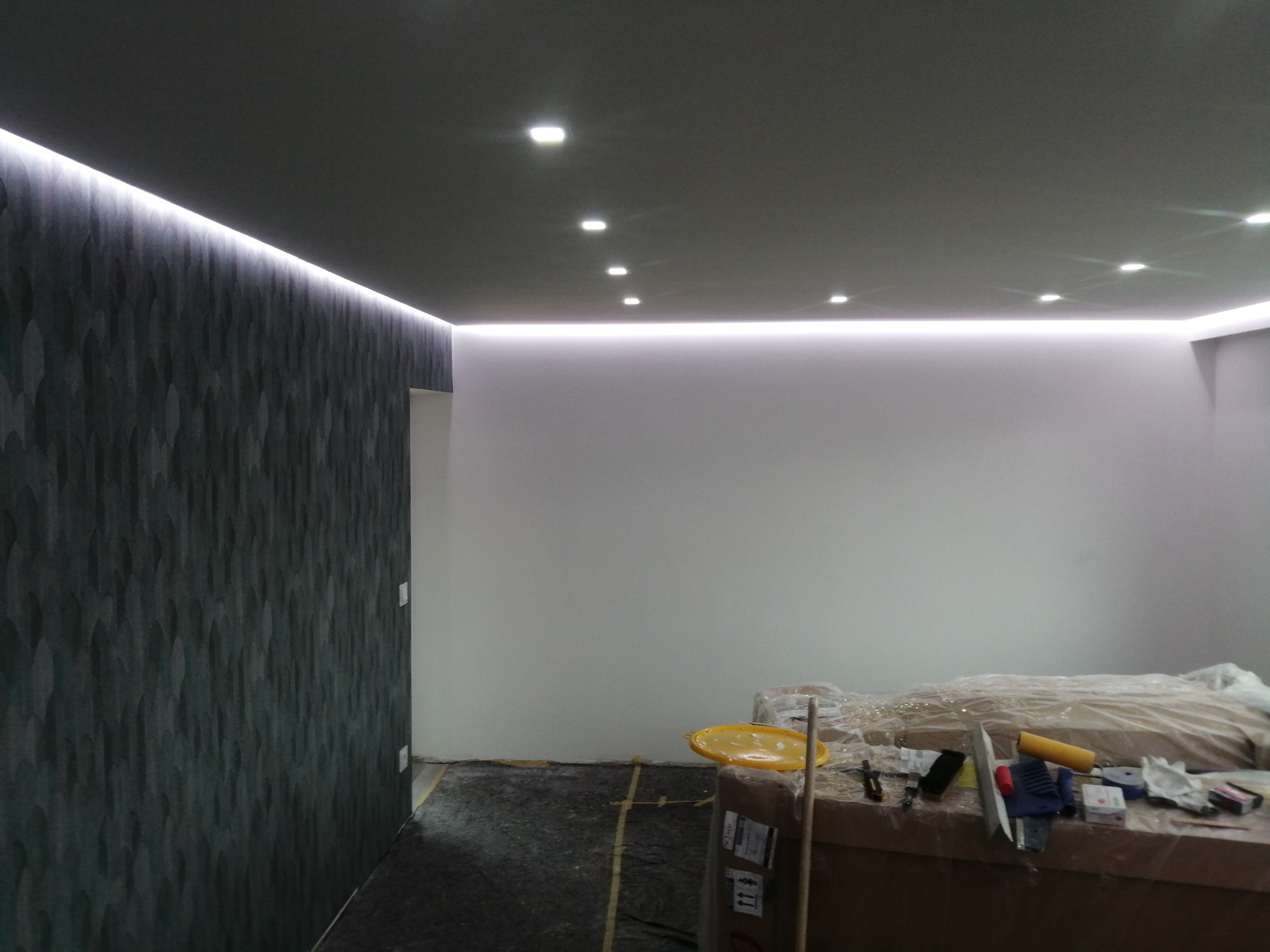 Nowoczesne wnętrze w trakcie wykańczania: ciemna ściana z fakturą, podwieszany sufit z oświetleniem LED, zabezpieczone meble i narzędzia malarskie na kartonach.