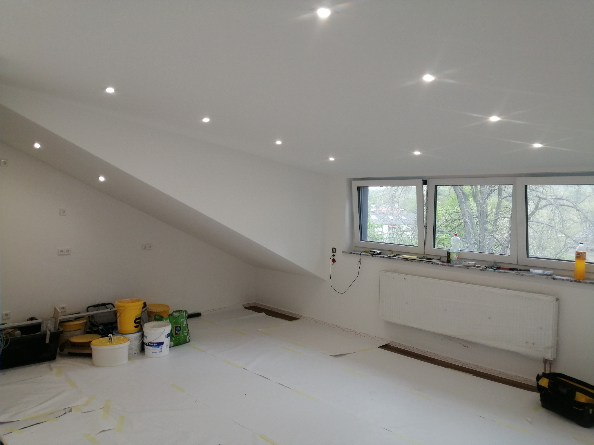Poddasze po malowaniu na biało, z widocznymi punktowymi lampami w suficie, zabezpieczoną podłogą folią malarską, wiadrami z farbą i narzędziami malarskimi przy ścianie szczytowej oraz zakrytym...