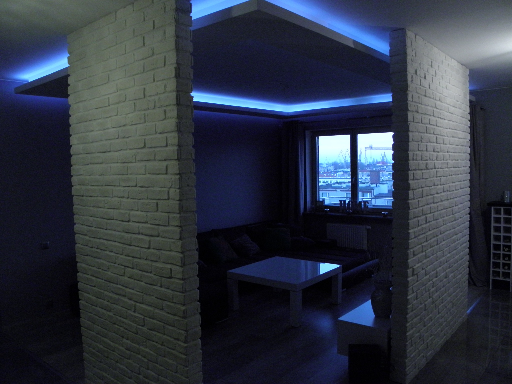 Nowoczesny salon z ceglanymi ściankami działowymi i podświetlanym sufitem w kolorze niebieskim, widok na miasto przez okno.