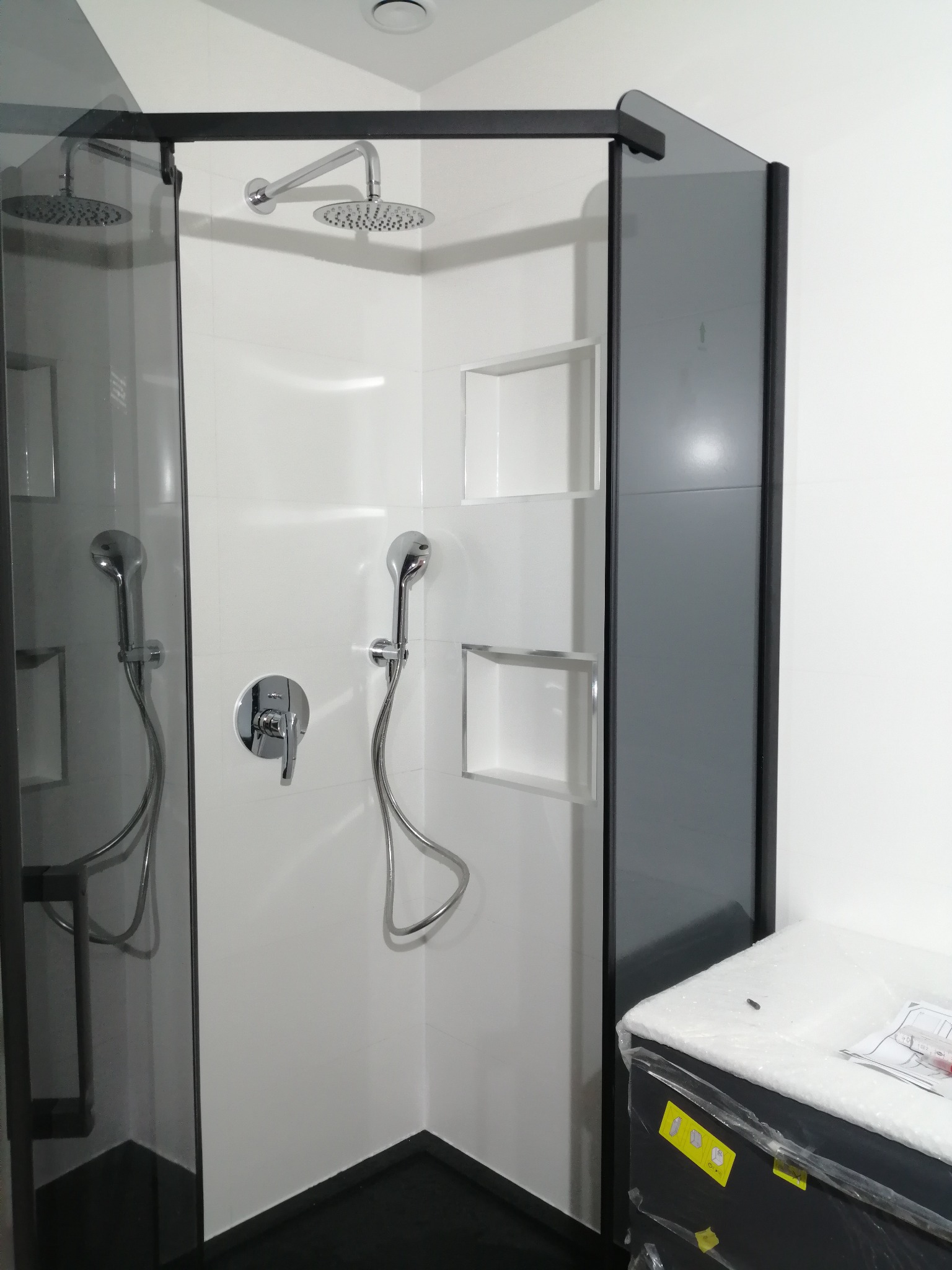 Nowoczesna kabina prysznicowa z czarnymi profilami, białymi płytkami i wnękami, wyposażona w deszczownicę i słuchawkę prysznicową. Częściowo zapakowana szafka obok.