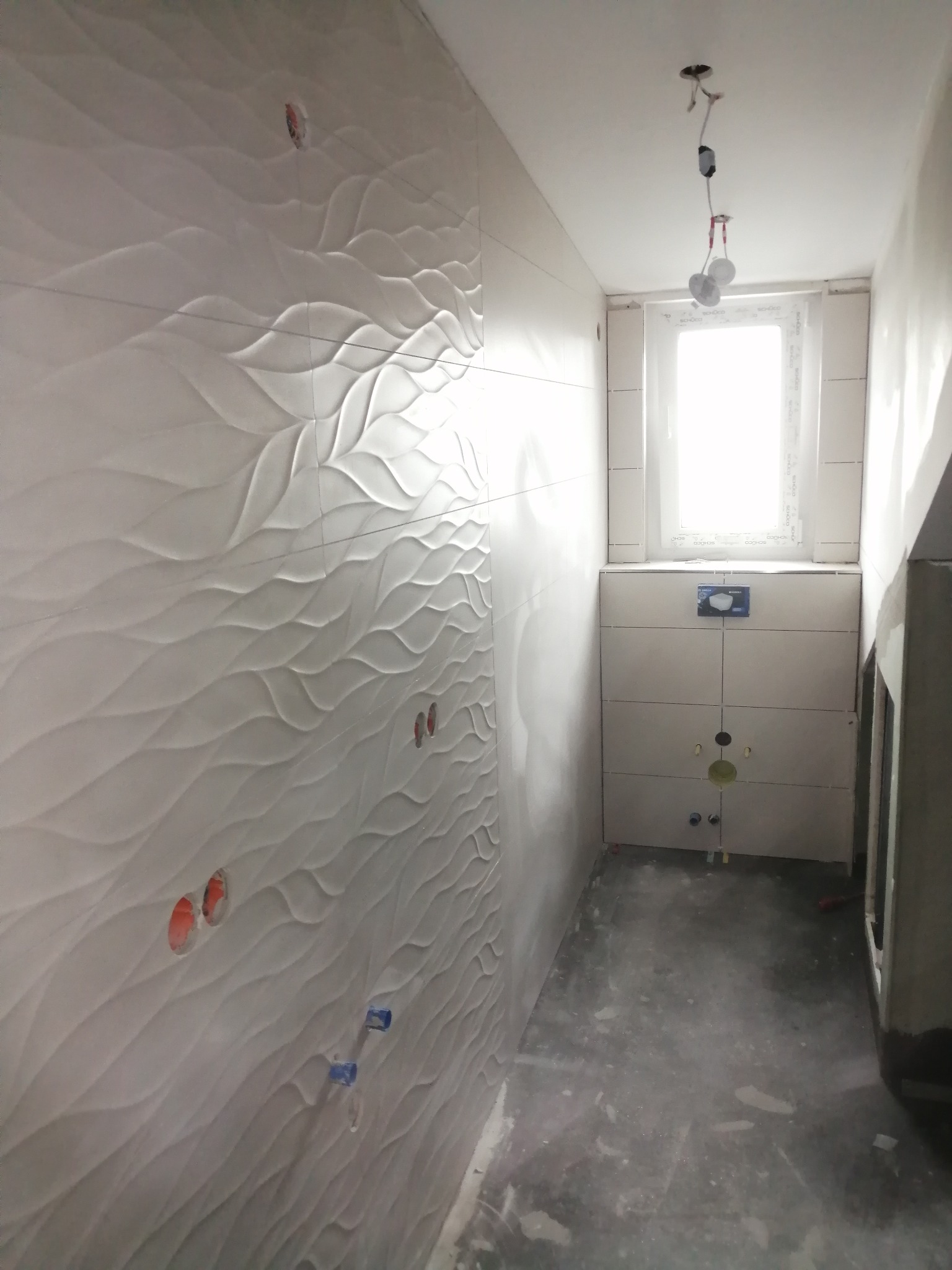 Łazienka w trakcie remontu w Szczecinie, z położoną białą, falistą glazurą na ścianie, widoczne otwory na instalacje i surowy beton na podłodze.