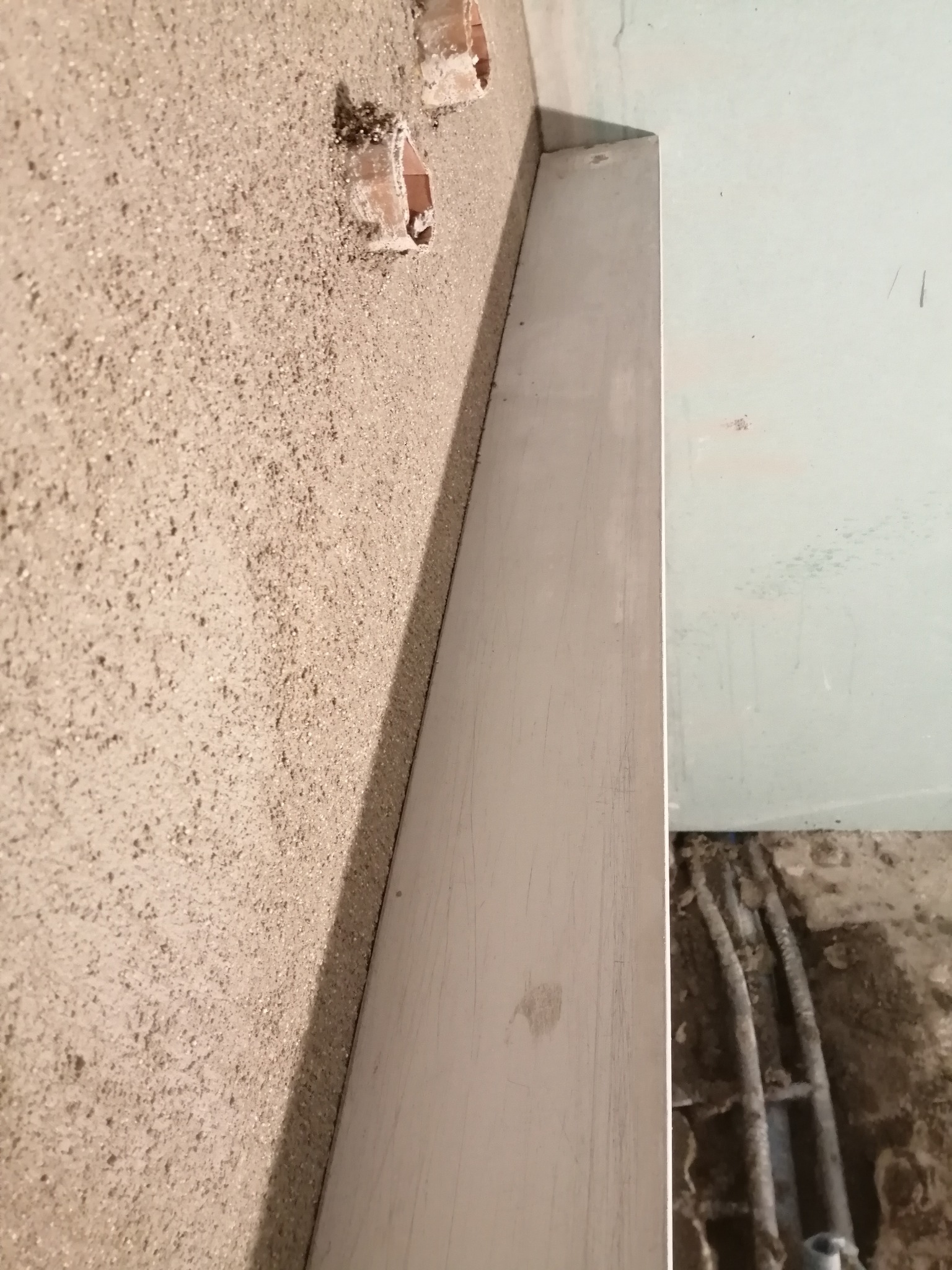 Ściana z nałożonym tynkiem strukturalnym, widoczne otwory pod instalacje i listwa przypodłogowa w trakcie montażu, fragment instalacji pod podłogą.
