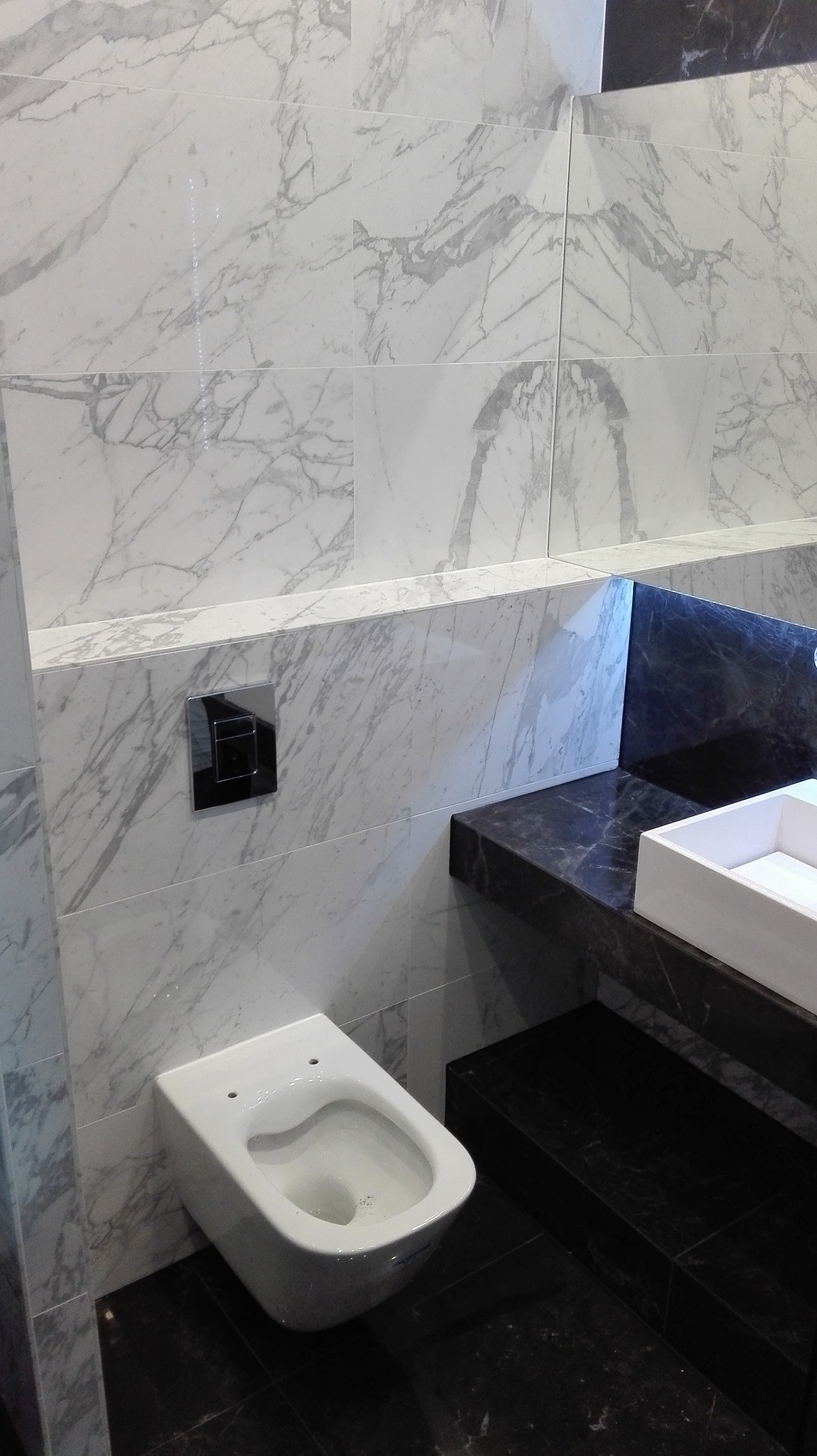 Nowoczesna toaleta podwieszana na tle ściany wyłożonej marmurowymi płytkami, z czarnym blatem i umywalką w tle. Widoczny chromowany przycisk spłuczki.