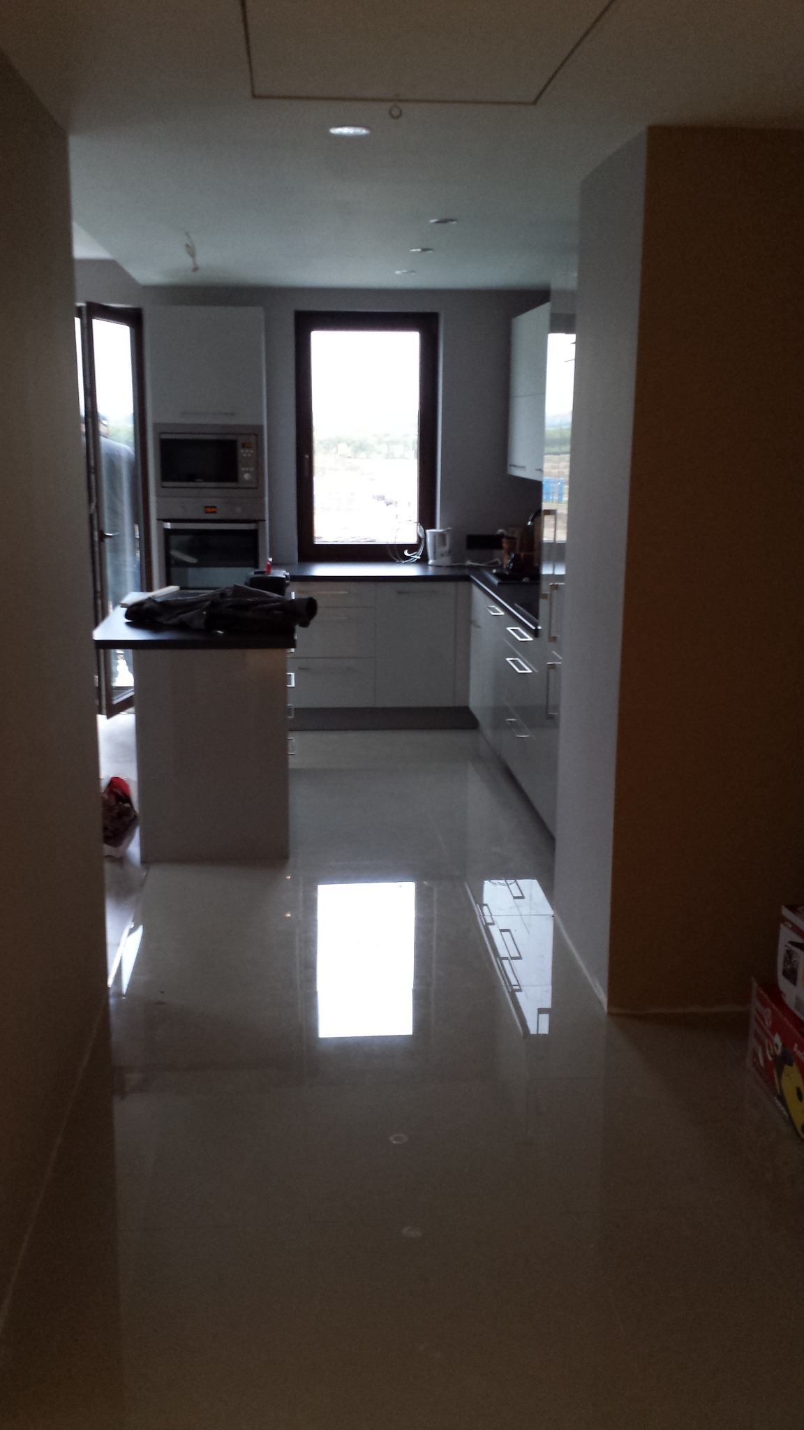 Nowoczesna, jasna kuchnia z błyszczącą, odbijającą światło podłogą. Widoczne białe szafki, wyspa kuchenna i okno. Na blacie leżą ubrania.