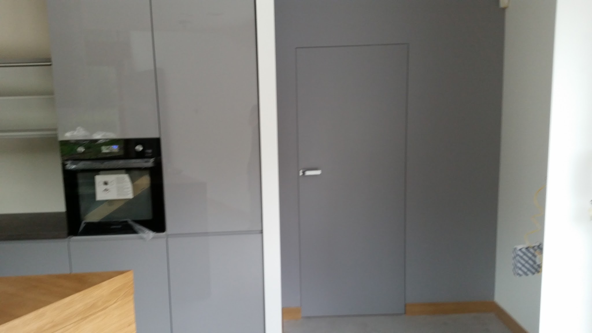 Nowoczesna kuchnia w szarościach z zabudowaną szafką z piekarnikiem i ukrytymi drzwiami, widoczne niedokończone prace elektryczne przy ścianie.