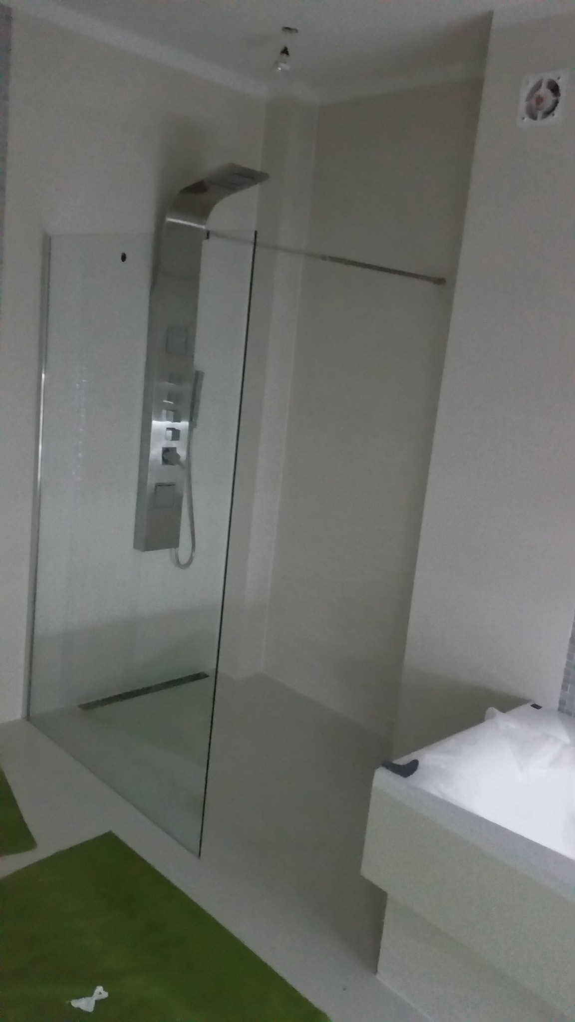 Nowoczesna kabina prysznicowa z panelem natryskowym ze stali nierdzewnej, widoczny odpływ liniowy, zielony dywanik i fragment wanny w minimalistycznej łazience wykończonej w jasnych odcieniach...