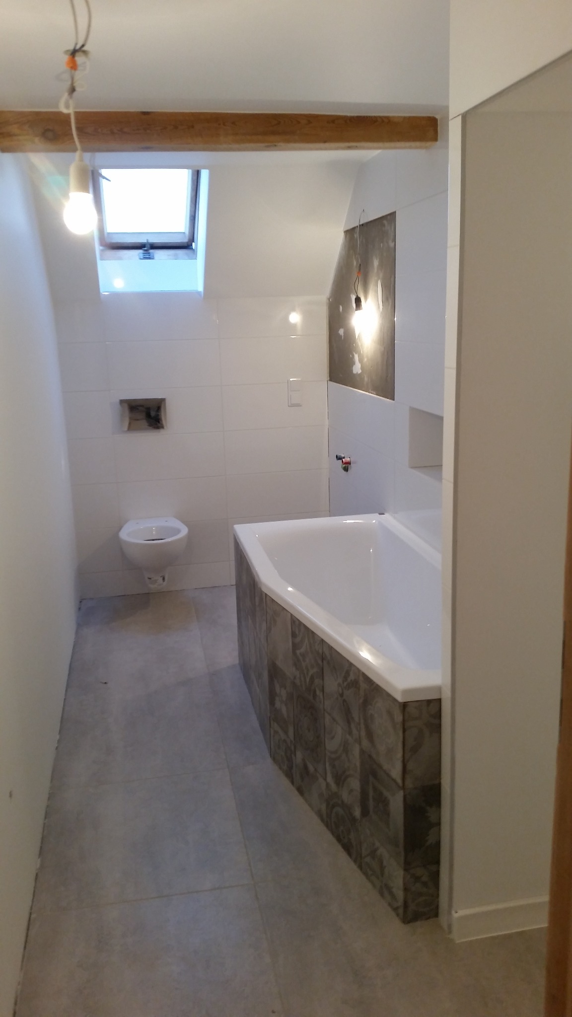 Łazienka w trakcie remontu: białe płytki na ścianach, częściowo obłożona płytkami wanna, podwieszana toaleta, okno dachowe i surowa belka drewniana z wiszącą żarówką.