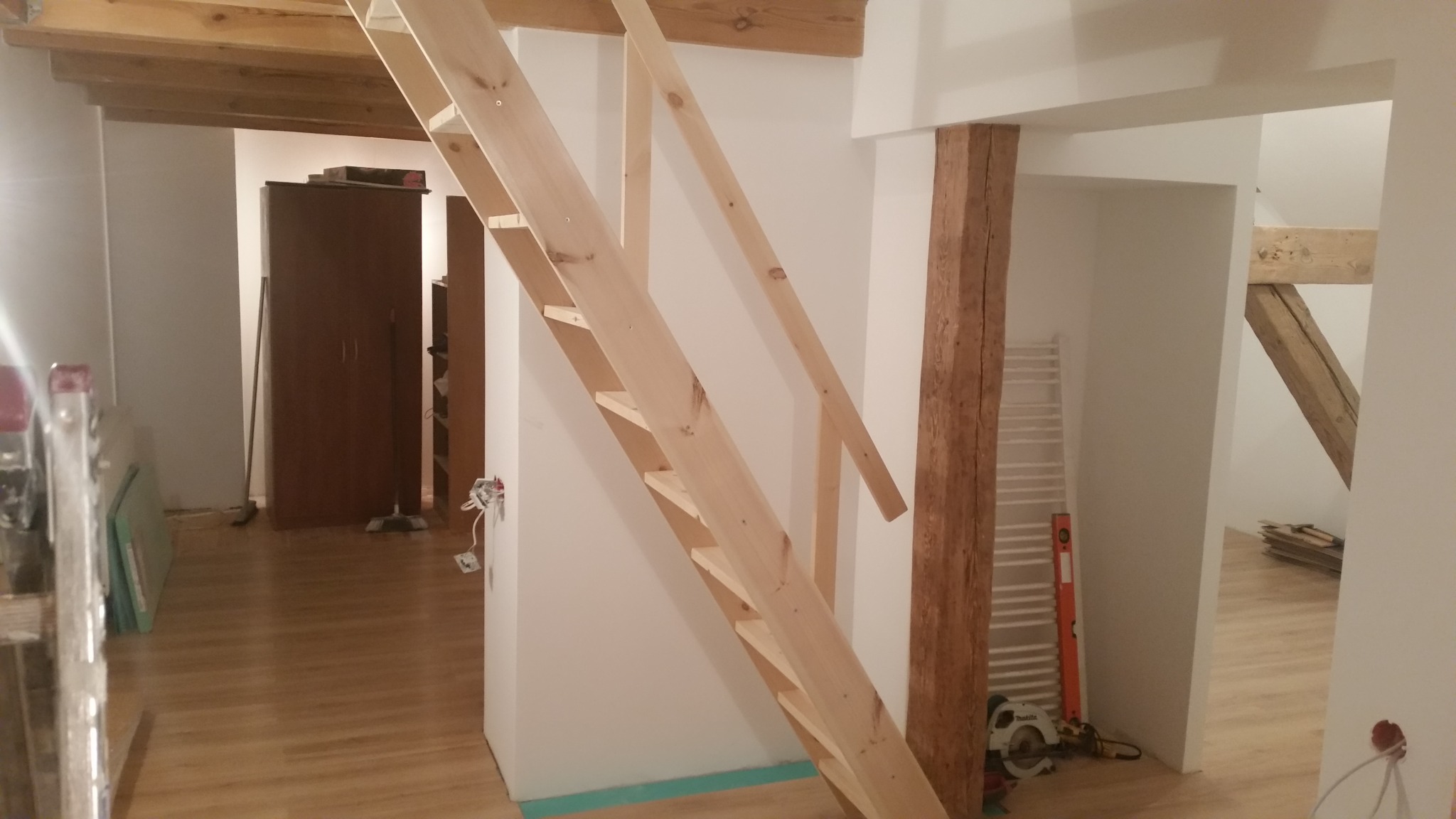 Wnętrze w trakcie remontu z drewnianymi schodami bez poręczy, widoczne odsłonięte przewody elektryczne i drewniana belka konstrukcyjna