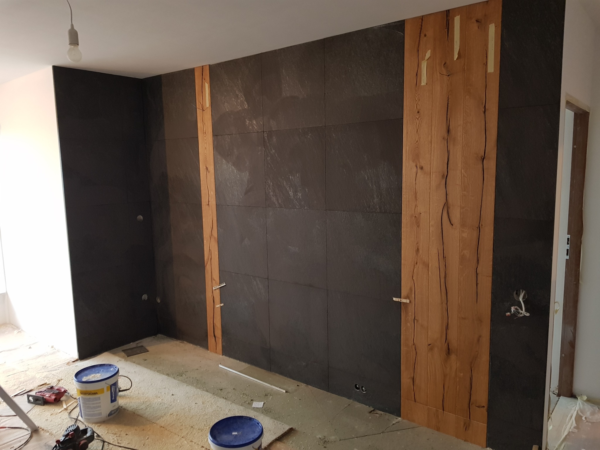 Ściana wyłożona ciemnymi płytkami z drewnianymi panelami po bokach, widoczne narzędzia i materiały budowlane na podłodze, wnętrze w trakcie wykończenia.