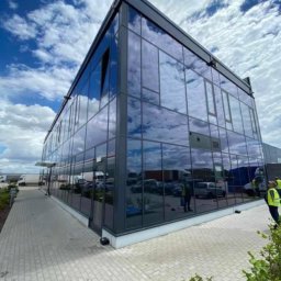 Kamar Service - Nowoczesny biurowiec z szklaną fasadą odbijającą błękitne niebo z chmurami. Widoczny chodnik z kostki brukowej i fragment zaparkowanych pojazdów.