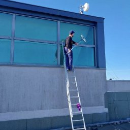 Kamar Service - Mężczyzna na drabinie myje okna budynku przemysłowego za pomocą ściągaczki. Widoczny fragment dachu, antena satelitarna i błękitne niebo.