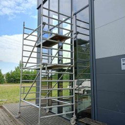 Kamar Service - Metalowe rusztowanie na kółkach ustawione przy szklanej fasadzie budynku. Widoczne odbicie otoczenia w szybach. Błękitne niebo z chmurami w tle.