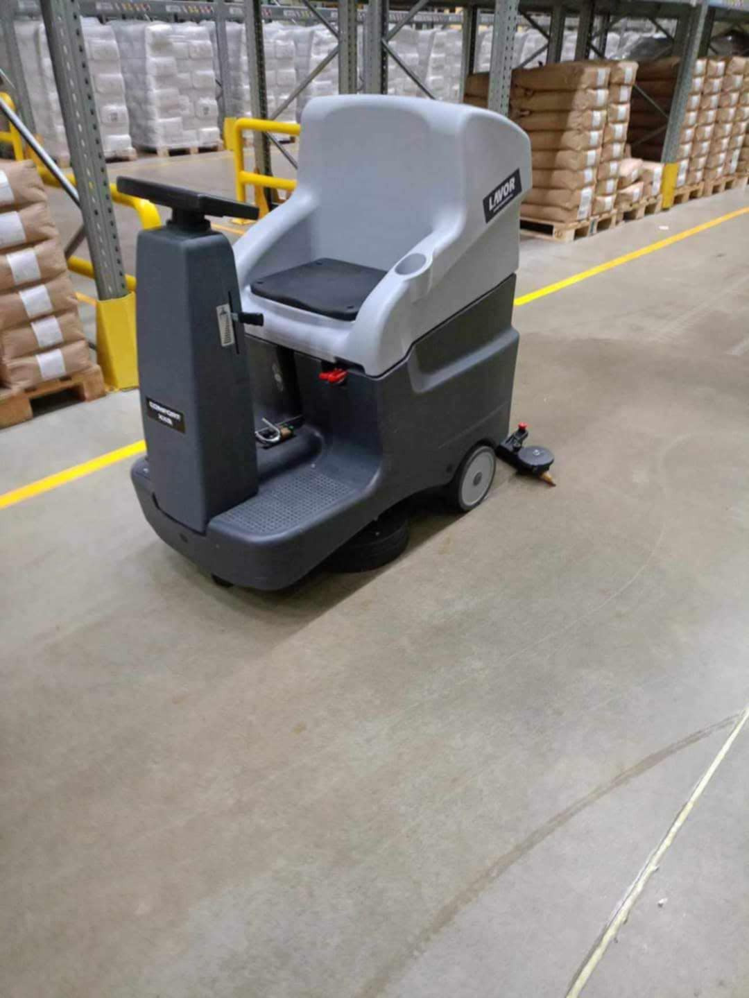 Samojezdna maszyna czyszcząca Lavor w magazynie, z widocznymi śladami po czyszczeniu na betonowej posadzce i paletami z towarem w tle. Żółte linie wyznaczają strefy.