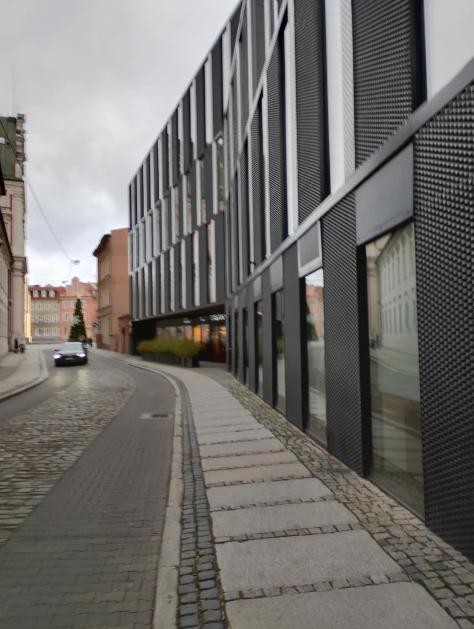 Ulica z kostki brukowej prowadząca obok nowoczesnego budynku biurowego z ciemną fasadą i dużymi oknami. W tle widoczny samochód i budynki w oddali.