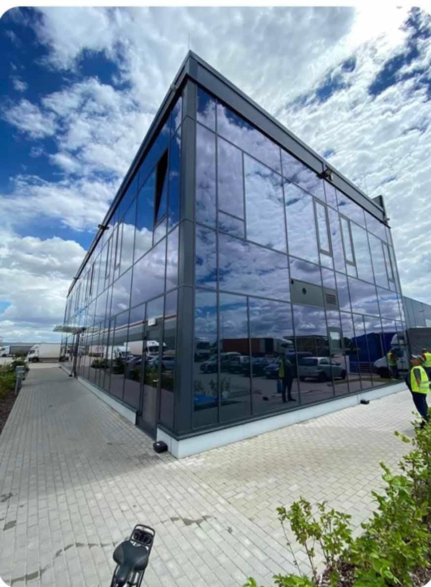 Nowoczesny biurowiec z szklaną fasadą odbijającą błękitne niebo z chmurami. Widoczny chodnik z kostki brukowej i fragment zaparkowanych pojazdów.