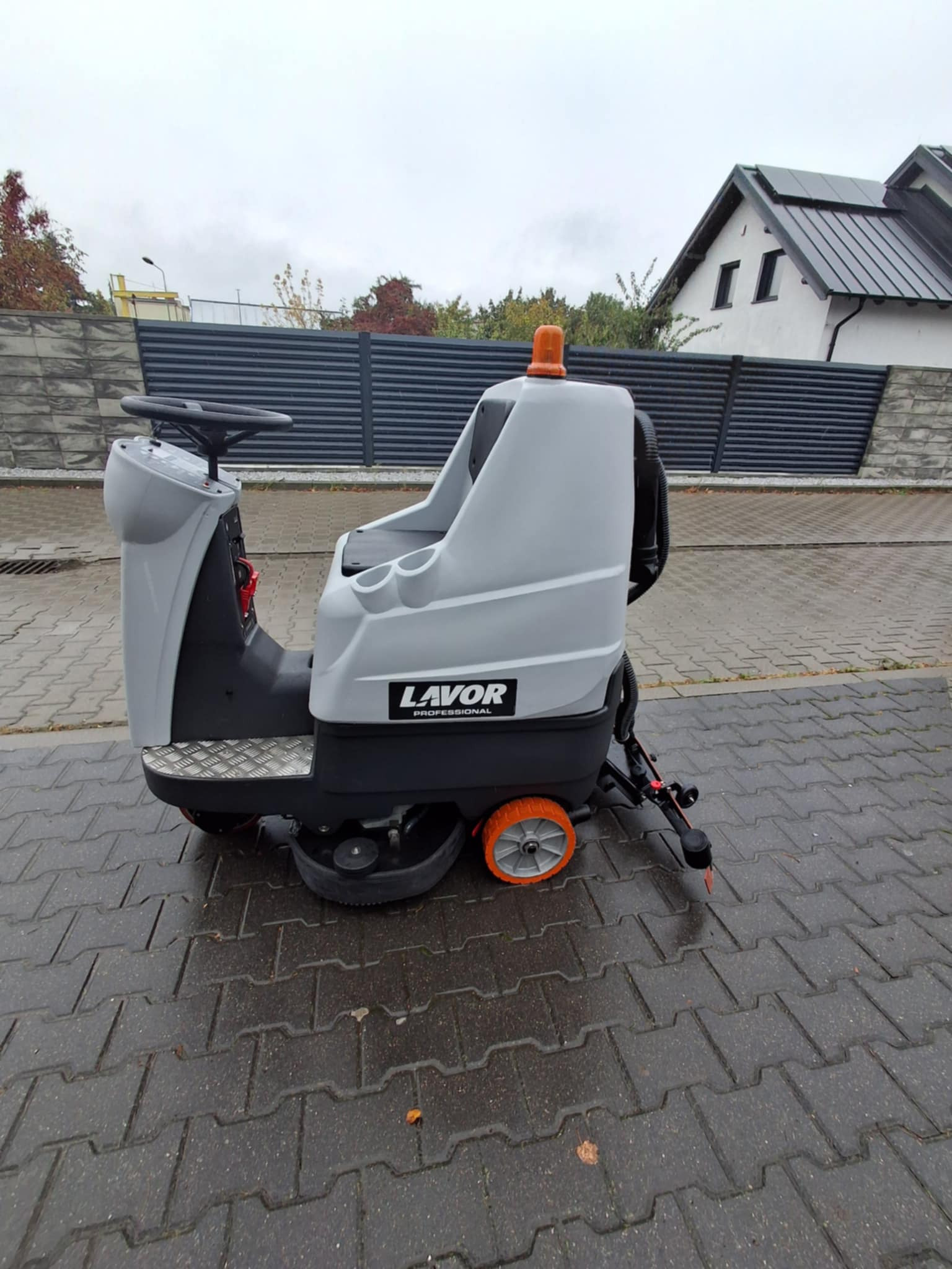 Profesjonalna, samojezdna maszyna czyszcząca Lavor na mokrej kostce brukowej. Widoczny budynek w tle. Pomarańczowe koła i szara obudowa.