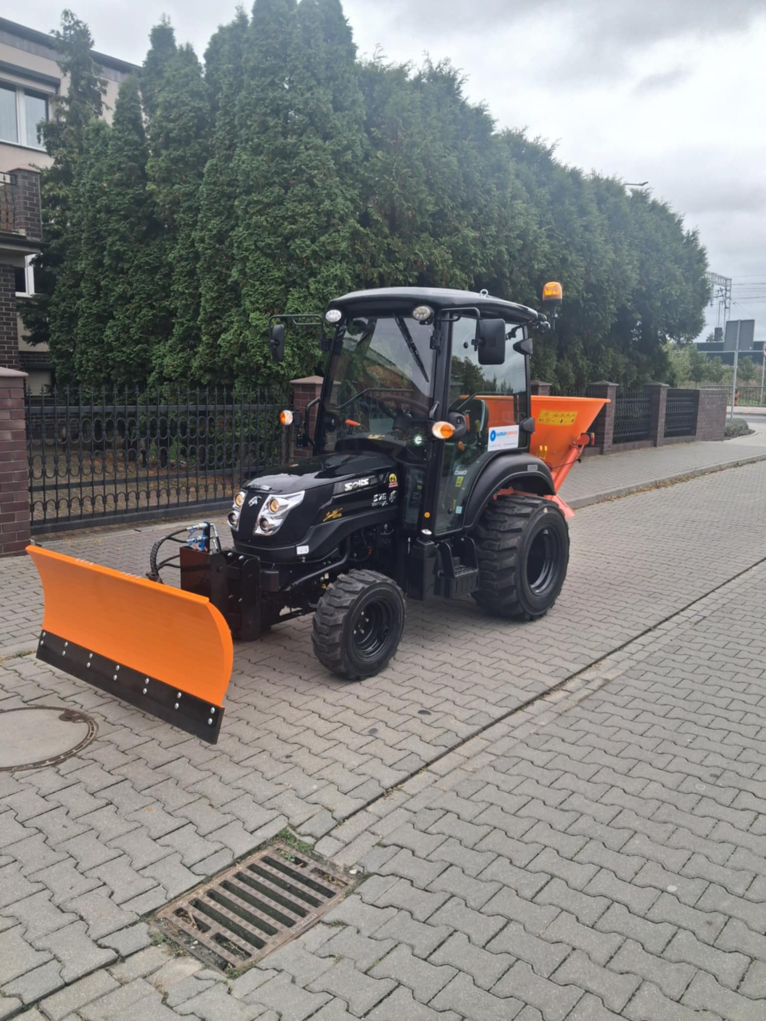 Czarny traktor z pomarańczowym pługiem i rozsiewaczem stoi na kostce brukowej. Widoczne budynki i drzewa w tle. Sprzęt gotowy do odśnieżania lub posypywania.