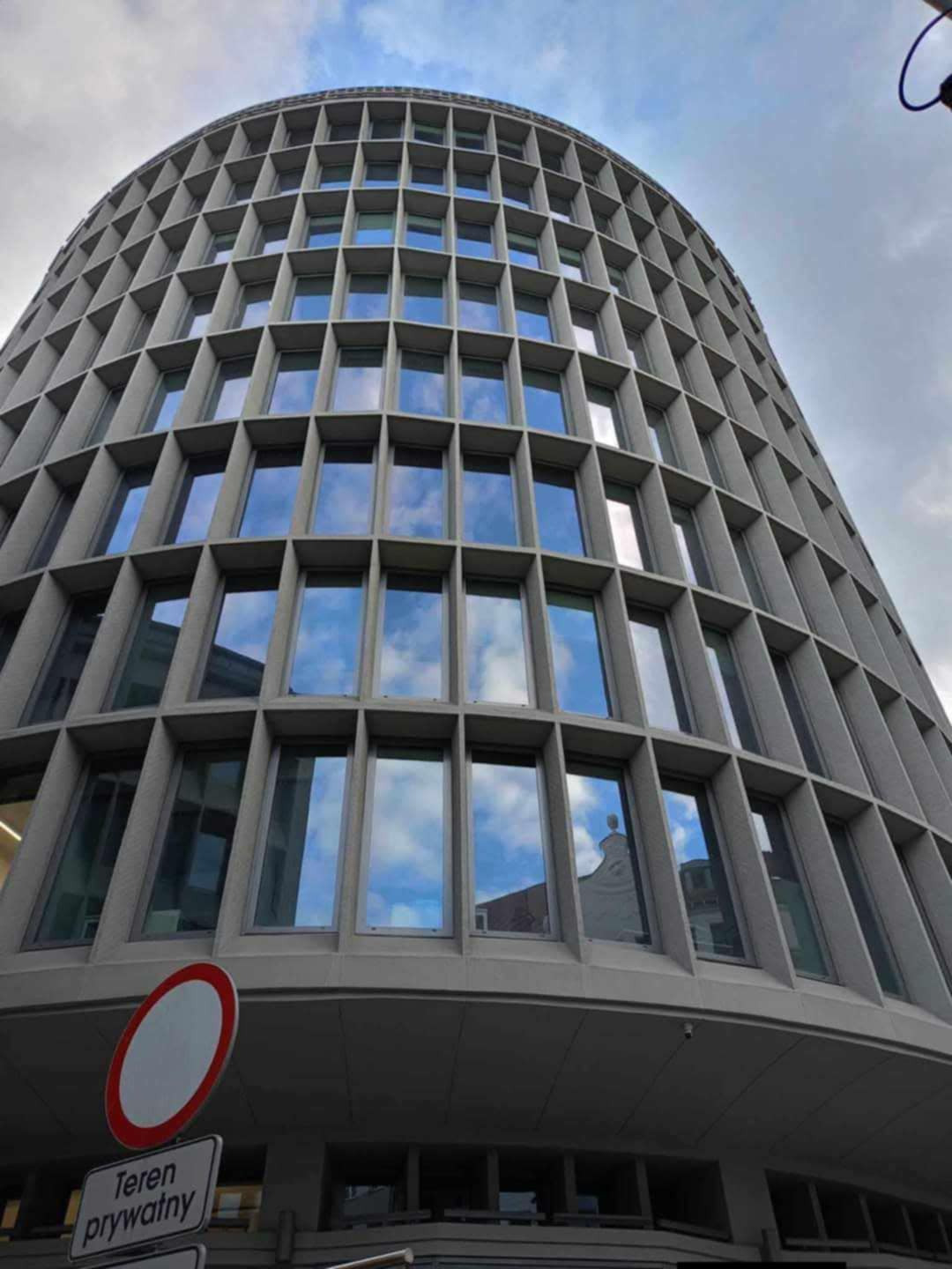 Wysoki, zaokrąglony budynek biurowy z licznymi oknami, w których odbija się niebo. Ujęcie z dołu, widoczny znak 'Teren prywatny'.