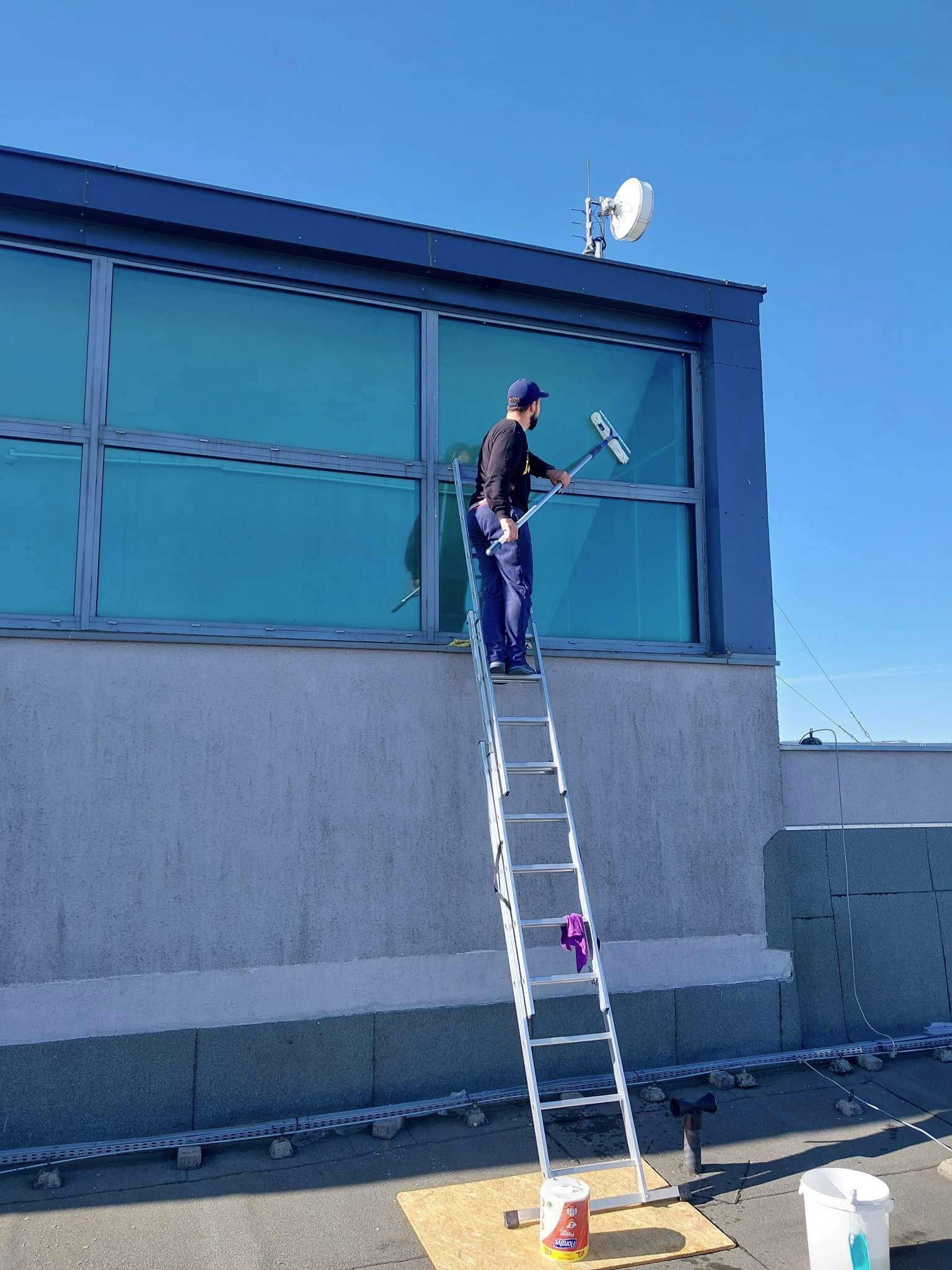 Mężczyzna na drabinie myje okna budynku przemysłowego za pomocą ściągaczki. Widoczny fragment dachu, antena satelitarna i błękitne niebo.
