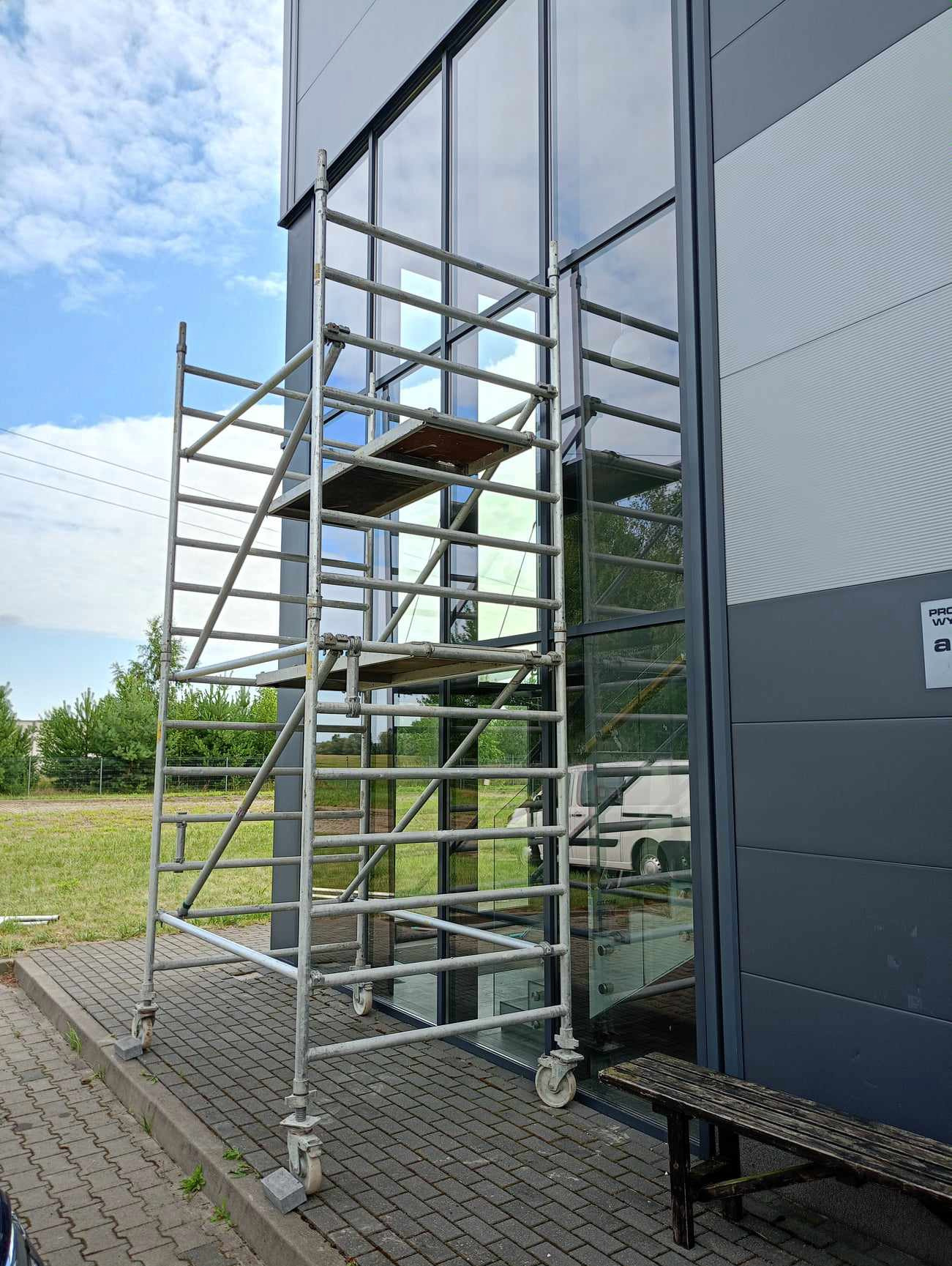 Metalowe rusztowanie na kółkach ustawione przy szklanej fasadzie budynku. Widoczne odbicie otoczenia w szybach. Błękitne niebo z chmurami w tle.
